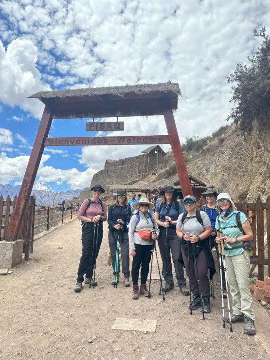 A group of hikers stands under a wooden entrance sign in Pisac, Peru, carrying walking poles and wearing outdoor gear. Ruins and a cloudy sky are visible in the background.