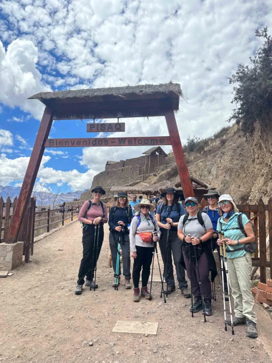 A group of hikers with trekking poles stands under a wooden sign reading "PISAC Bienvenidos - Welcome," with ancient ruins in the background.