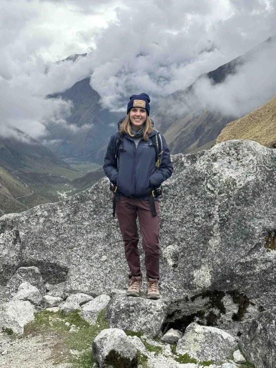 A person stands on rocky terrain wearing hiking gear, with a cloudy mountain landscape in the background.