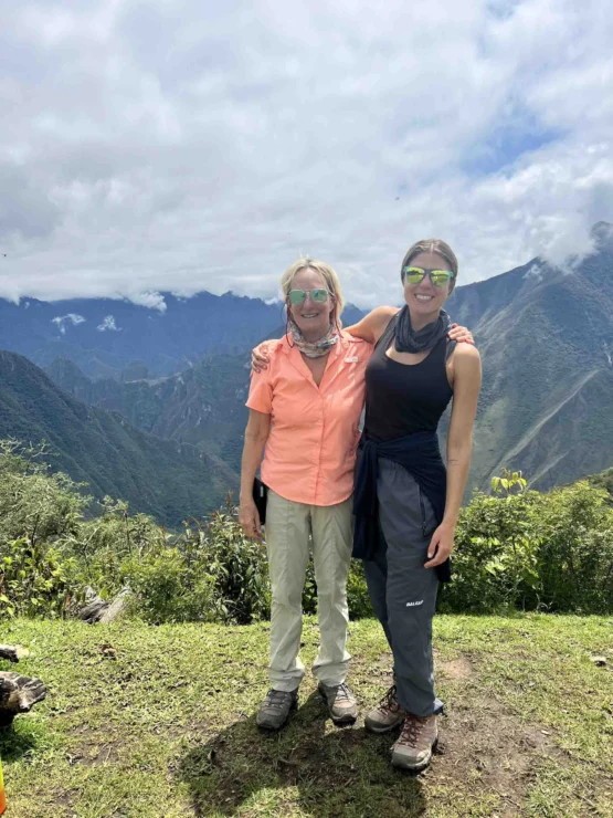 Two people in outdoor gear stand on a grassy area with a mountainous landscape in the background. Both are smiling and wearing sunglasses.