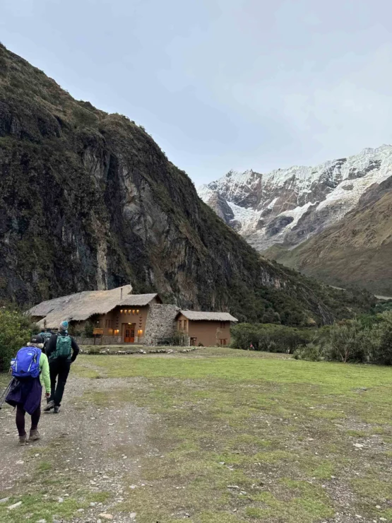 Two hikers walk toward a rustic building, surrounded by grassy terrain and towering mountains with snow-covered peaks in the background.