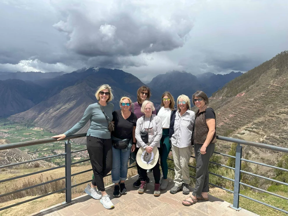 A group of seven people stands on a viewing platform overlooking a mountainous landscape under a cloudy sky.