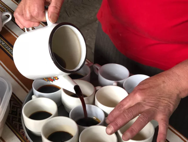 A person in a red shirt pours coffee from a white jug into multiple white cups arranged on a tray.