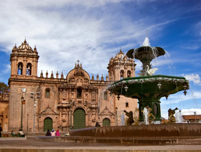 A historic cathedral with twin bell towers stands behind a large ornate fountain in a sunny plaza.