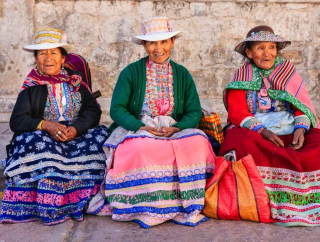 Three women in traditional colorful clothing and hats sit outdoors against a stone wall, with woven bags beside them.