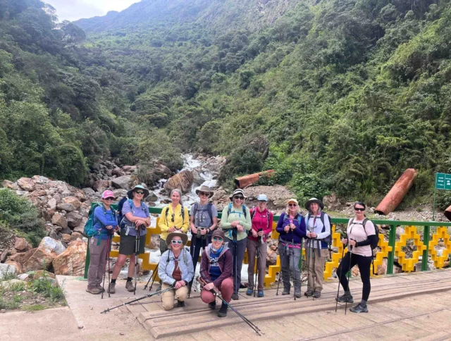 A group of hikers with trekking poles pose on a bridge surrounded by lush green mountains and a flowing stream.