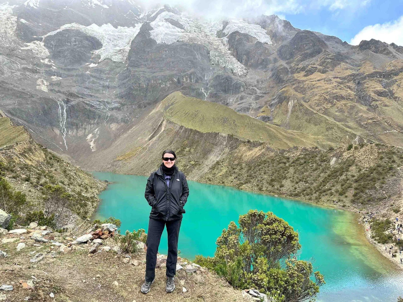 Person standing in front of a vibrant turquoise lake surrounded by mountainous terrain under a cloudy sky.