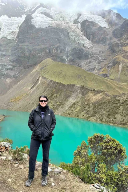 Person standing in front of a vibrant turquoise lake surrounded by mountainous terrain under a cloudy sky.