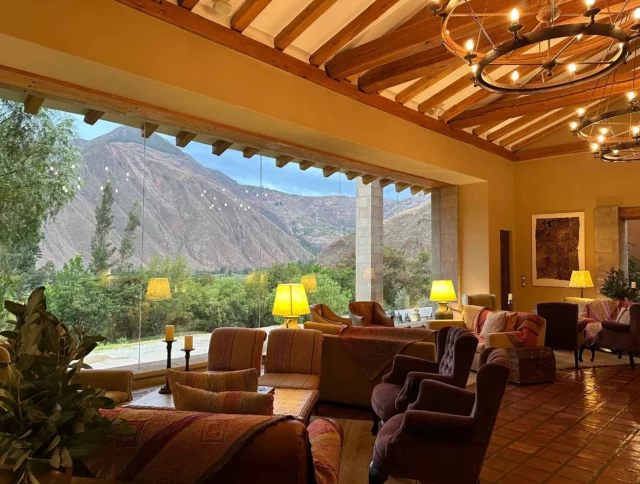 Lounge area with large windows showing a scenic mountain view, warm lighting from lamps and chandeliers, and comfortable seating with cushions and blankets.