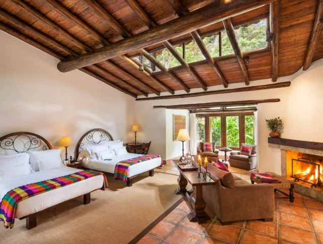 A cozy room with two beds, colorful blankets, a fireplace, chairs, and a large window with a garden view. The ceiling features exposed wooden beams.