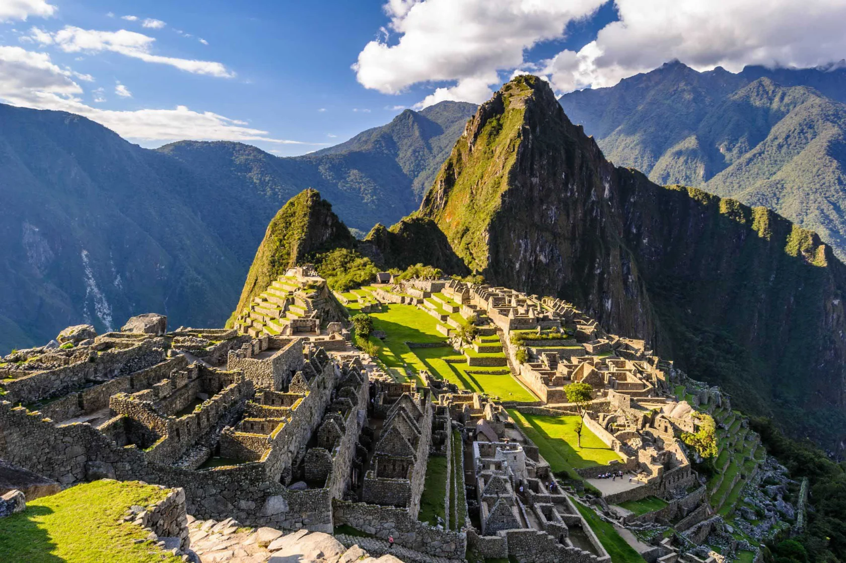 Aerial view of Machu Picchu, showcasing ancient stone structures and terraces set against a backdrop of lush mountains and blue sky.