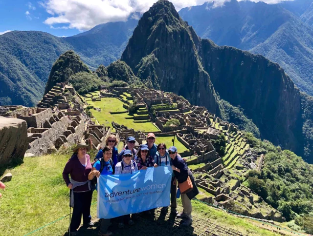 A group of women holding a banner stand in front of the historic ruins of Machu Picchu, surrounded by mountains under a clear blue sky.