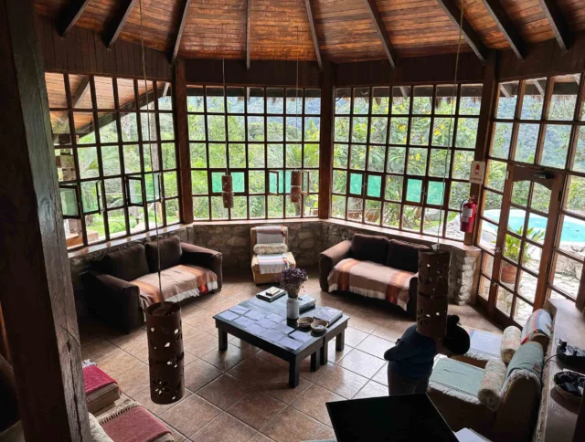 A rustic room with large windows, wooden ceiling, and stone walls. Sofas and a central coffee table furnish the space, overlooking a green landscape and a pool.