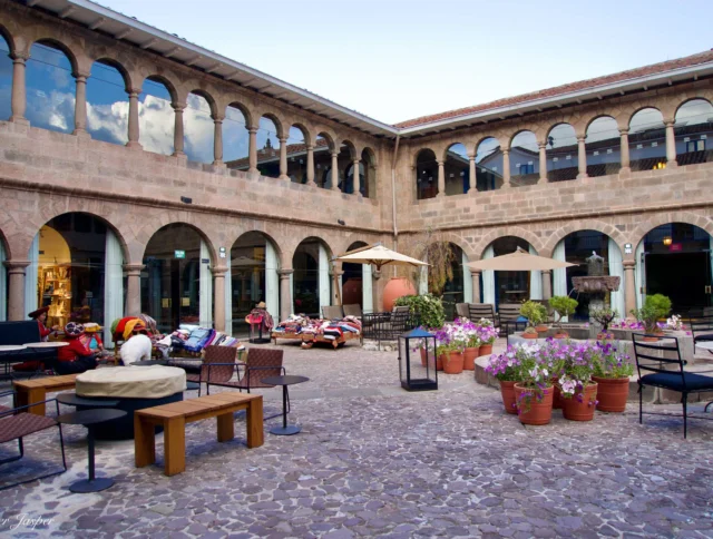 A courtyard with stone flooring, surrounded by an arched building. There are tables, chairs, potted flowers, and a few colorful textiles on display.