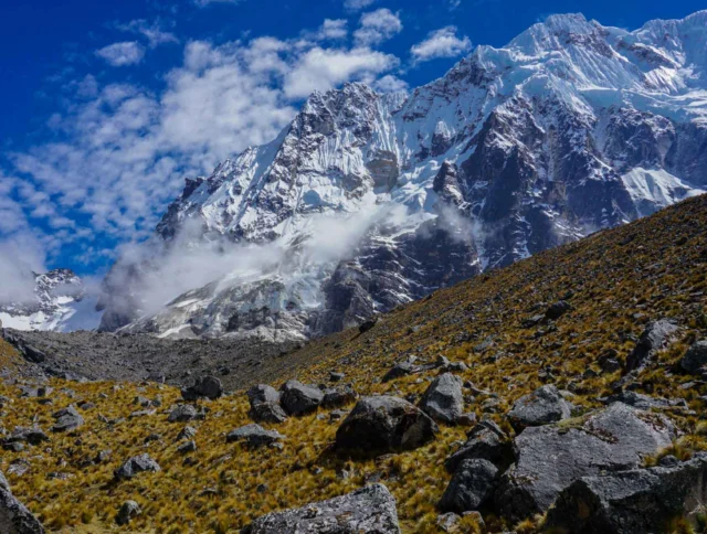 A rugged mountain landscape with snowy peaks under a blue sky, scattered clouds, and a rocky foreground.