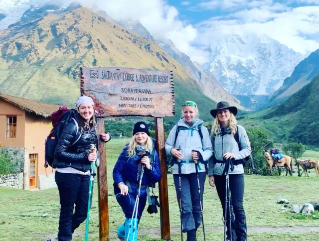 Four hikers pose with trekking poles in front of a wooden sign at Salkantay Lodge & Adventure Resort. Snow-capped mountains are visible in the background.