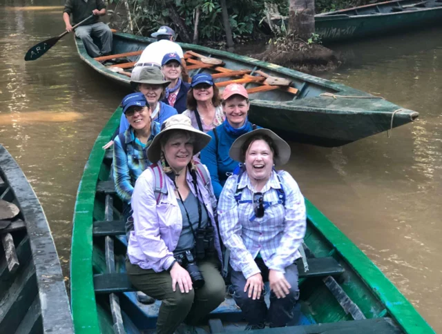 A group of people in hats sitting in a green canoe on a river surrounded by dense vegetation.