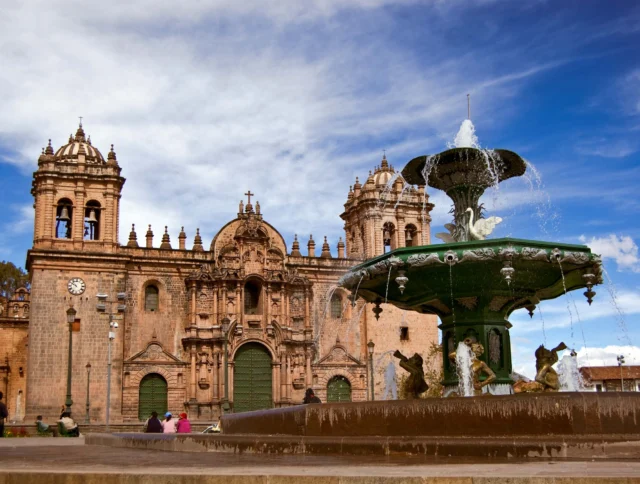 Fountain in front of a historic church with two bell towers under a blue sky with clouds.