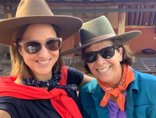 Two people wearing sunglasses, hats, and bandanas smile for a selfie outdoors.