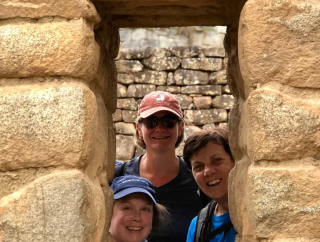 Three people smiling through a stone window at an ancient ruin site.