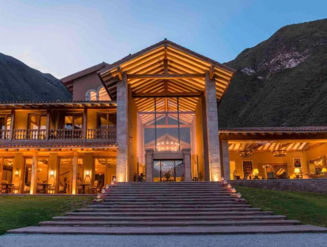 A large, lit-up lodge with a steep staircase, surrounded by mountains in a twilight setting. The entrance features tall glass doors and rustic wooden beams.