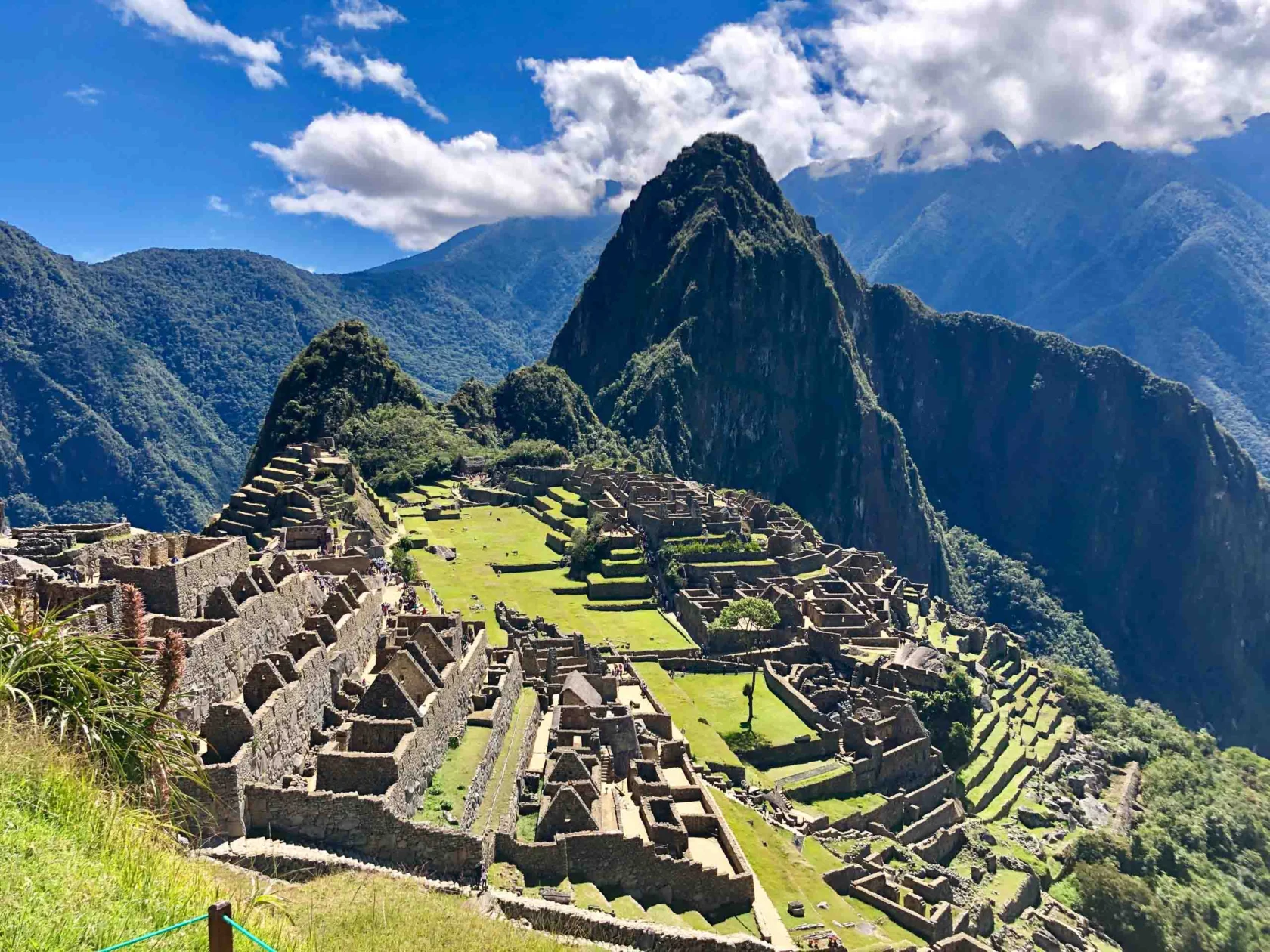 Sunny view of Machu Picchu ruins with terraced slopes, stone structures, and towering mountains in the background under a partly cloudy sky.