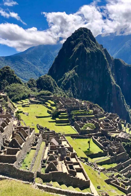 Sunny view of Machu Picchu ruins with terraced slopes, stone structures, and towering mountains in the background under a partly cloudy sky.