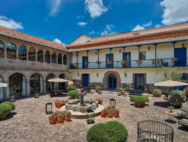 A historic courtyard with stone flooring, a central fountain, and potted plants. The double-story building has arches and blue balconies, and the sky is clear with a few clouds.