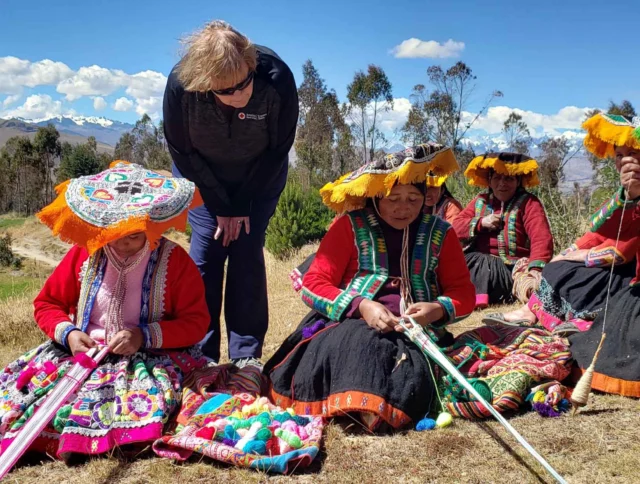 A person observes four women in colorful traditional attire weaving outdoors in a mountainous landscape.