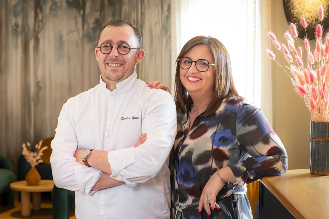 A smiling man in a white chef's coat stands with a woman in a patterned blouse. They are indoors with decorative elements in the background.