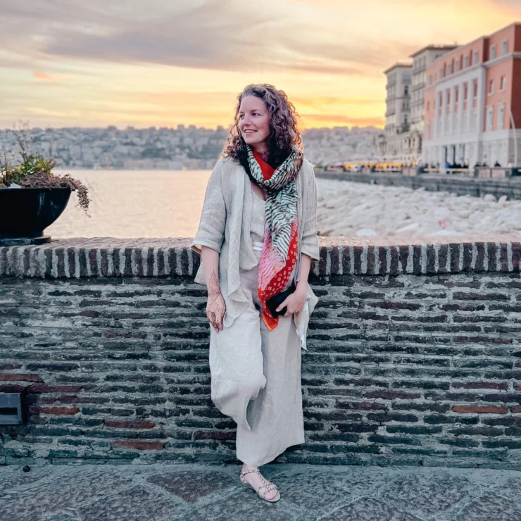 A person stands against a brick wall, wearing a scarf and light clothing, with a sunset and cityscape in the background.