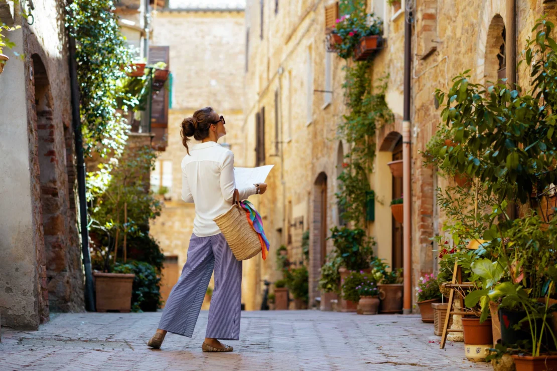 Woman in a white shirt and striped pants holds a map and stands in a narrow, historic alleyway with stone buildings and plants.