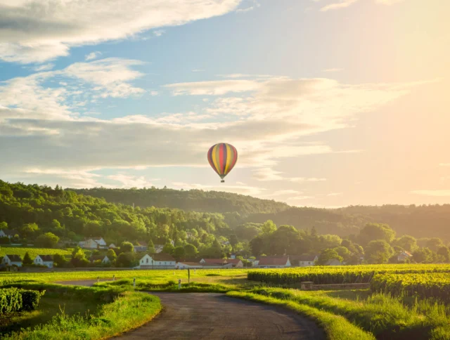 A hot air balloon floats over a rural landscape with fields, a winding road, and a small village under a partly cloudy sky at sunset.