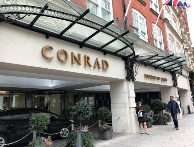 Entrance of Conrad London St. James hotel, featuring a car, potted plants, and two people walking by under a glass canopy.