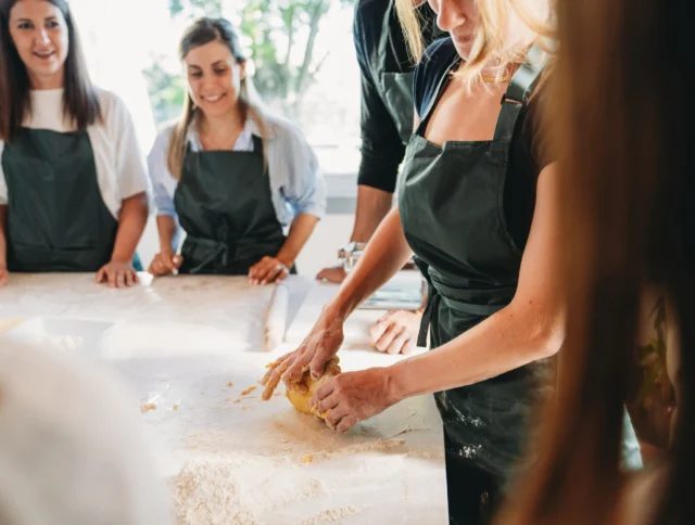 A group of people wearing aprons are gathered around a table, watching someone knead dough.