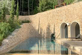 Stone wall with arched openings and a small, clear swimming pool in a wooded area.