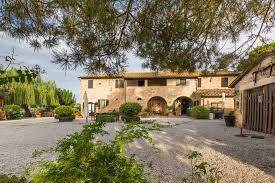 A rustic stone building with arched entrances, surrounded by greenery and a gravel courtyard under a cloudy sky.