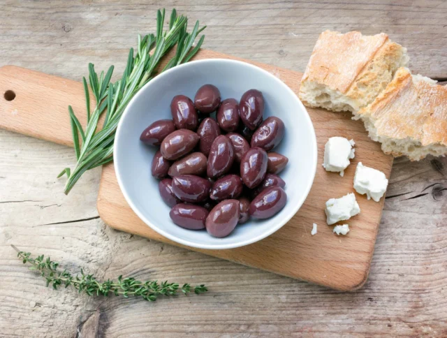 A bowl of black olives on a wooden board, accompanied by rosemary, thyme, two pieces of bread, and cubes of cheese.