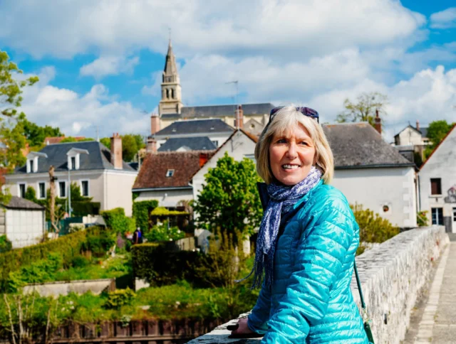 A woman in a blue jacket stands on a stone bridge, with a scenic village and church in the background under a partly cloudy sky.