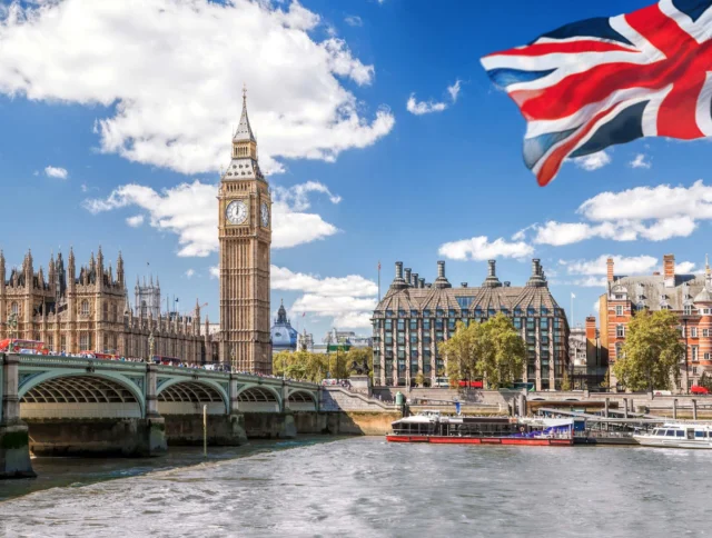 Big Ben and the Houses of Parliament beside the River Thames in London, with a Union Jack flag in the foreground and a boat on the water.