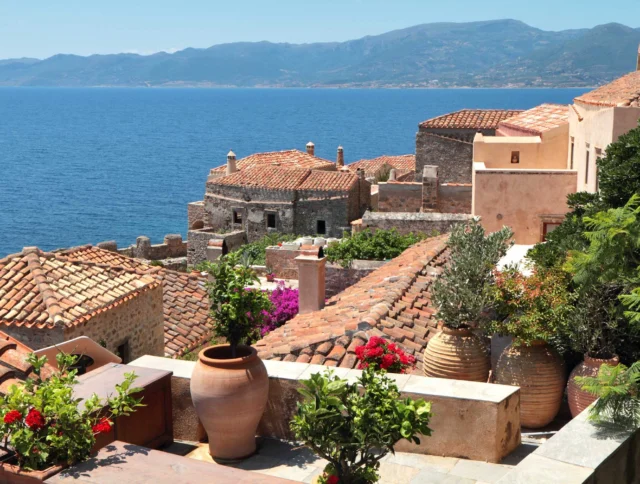 Mediterranean seaside village with terracotta-roofed stone buildings, potted plants, and a view of a calm blue sea against a mountainous backdrop.