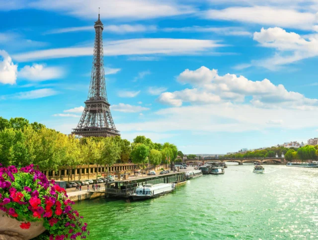 The Eiffel Tower by the Seine River with boats and trees, under a blue sky with clouds and a foreground of colorful flowers.