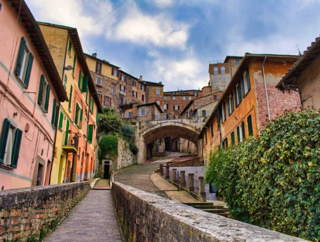 A narrow cobblestone path leads through a picturesque street with colorful buildings and an arched stone bridge under a cloudy sky.