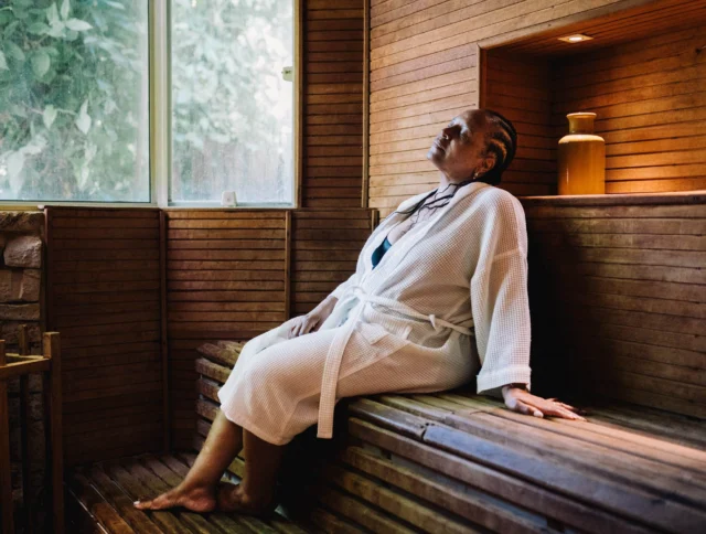 A person wearing a robe relaxes on a wooden bench inside a sauna, with natural light coming through a window.