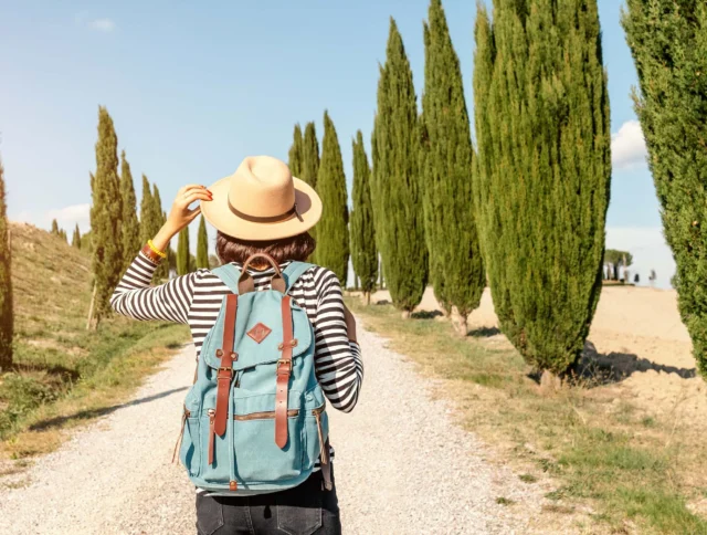 Person with a striped shirt and blue backpack walks down a tree-lined gravel path, holding a straw hat.