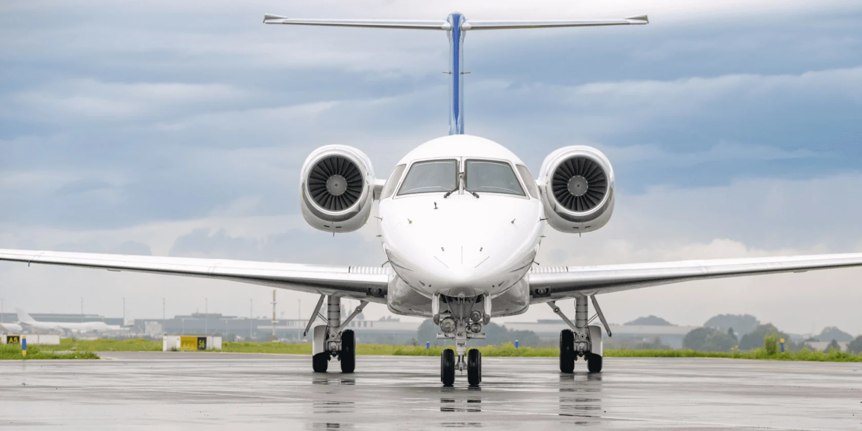 Front view of a white private jet on a wet runway, with a cloudy sky in the background.