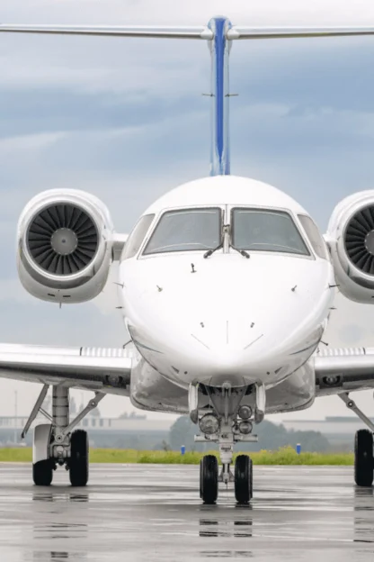 Front view of a white private jet on a wet runway, with a cloudy sky in the background.