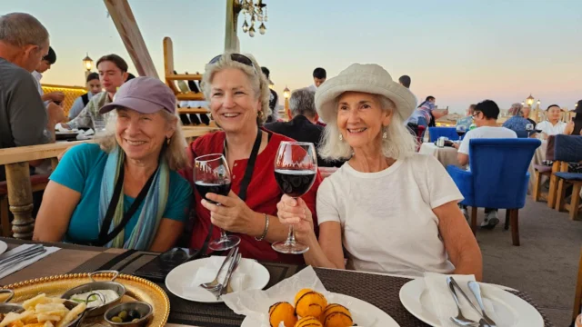 Three women smiling and holding glasses of red wine at an outdoor dining area, with plates of food on the table.