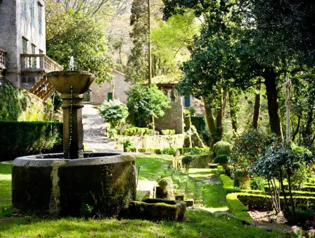 Lush garden with a stone fountain, manicured hedges, and vibrant green foliage under dappled sunlight. Stone paths and walls are visible in the background.