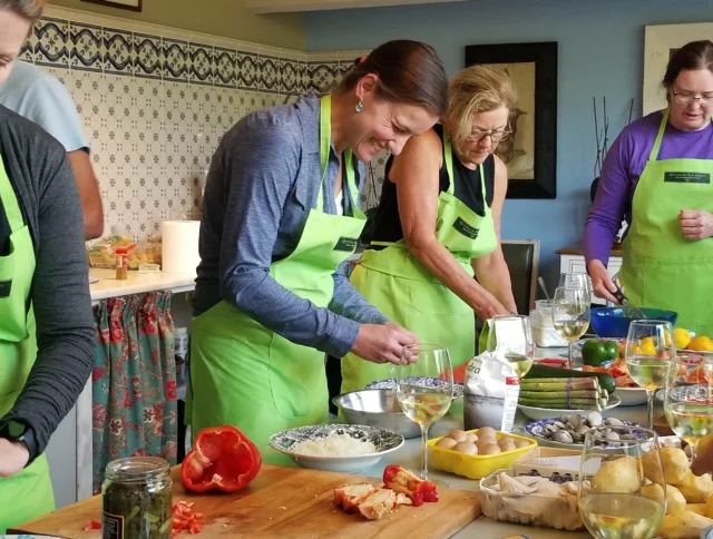 People wearing green aprons are preparing food in a kitchen, with various ingredients and glasses of white wine on the counter.